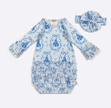 A baby long-sleeve romper in white and blue with a repeating castle and bow toile pattern, shown with a matching fabric headband on a light gray background.