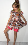A young girl with blonde hair and a brown bow headband is wearing a tiered ruffle dress with a brown, black, and white print featuring illustrations of hot air balloons, with bright pink ruffle trim. She is posing against a plain white wall.