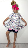 A young girl with a blonde bun and a large pink bow headband models a short-sleeve white tunic top with a navy blue and pink floral/dog pattern, paired with denim shorts and a pink gingham headband, posing against a white wall.