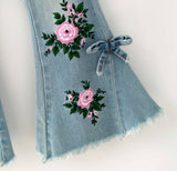 Light blue denim bell-bottom jeans with floral embroidery, featuring pink roses and green leaves, a denim bow detail, and raw frayed hems.
