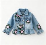 A light blue denim jacket for toddlers or girls, decorated with pink and green floral embroidery, bow details on the chest pockets, and ruffled cuffs and hem, shown flat on a white background.