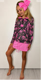 A young girl modeling a long-sleeve collared shirt with a pink and black camouflage/tree-branch print, paired with a bright pink mini skirt and a large pink bow headband, standing against a plain light wall.