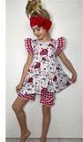 A young girl modeling a red and white gingham and barn print dress with matching ruffle shorts and a red bow headband, standing against a plain white wall.