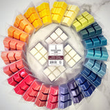 Colorful wax melts in clear packaging arranged in a circular pattern on a white surface.