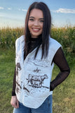 A woman wearing a light gray zippered vest with black cow and rider illustrations, layered over a black long-sleeve top and blue jeans, standing outdoors in a grassy field under a blue sky.