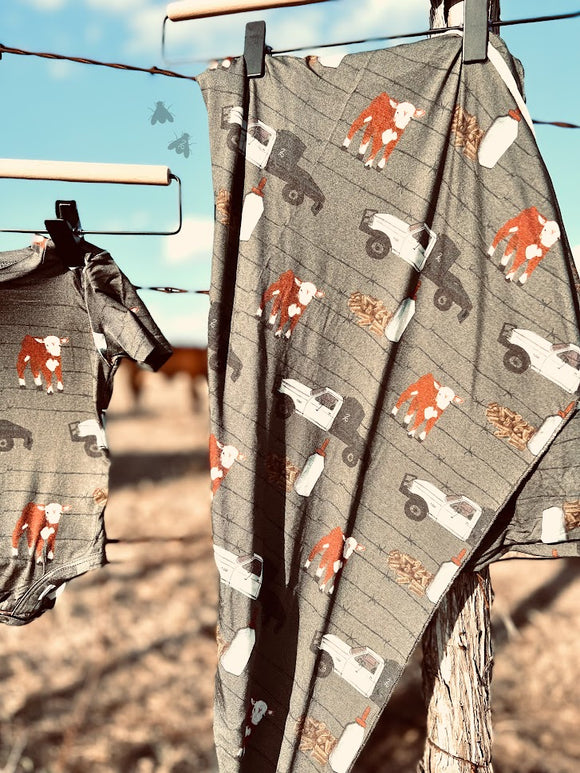 Swaddle blanket with ranch design hanging on a barbed wire fence next to a matching onesie. Pattern has hereford cow, truck, barbed wire, and bottle 2 Fly Co 